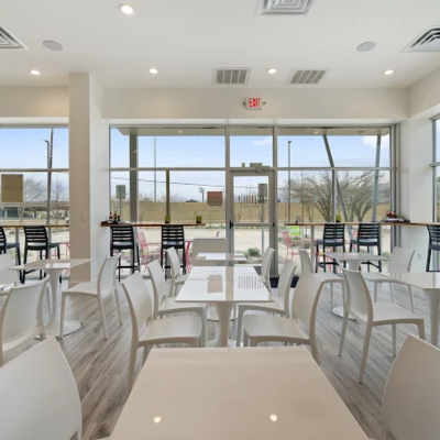 Bright cafe with white tables, modern chairs, bar-height counters, and large floor-to-ceiling windows.